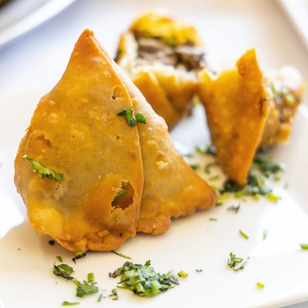 Golden-brown Indian samosas, garnished with fresh green herbs, are presented on a white plate.
